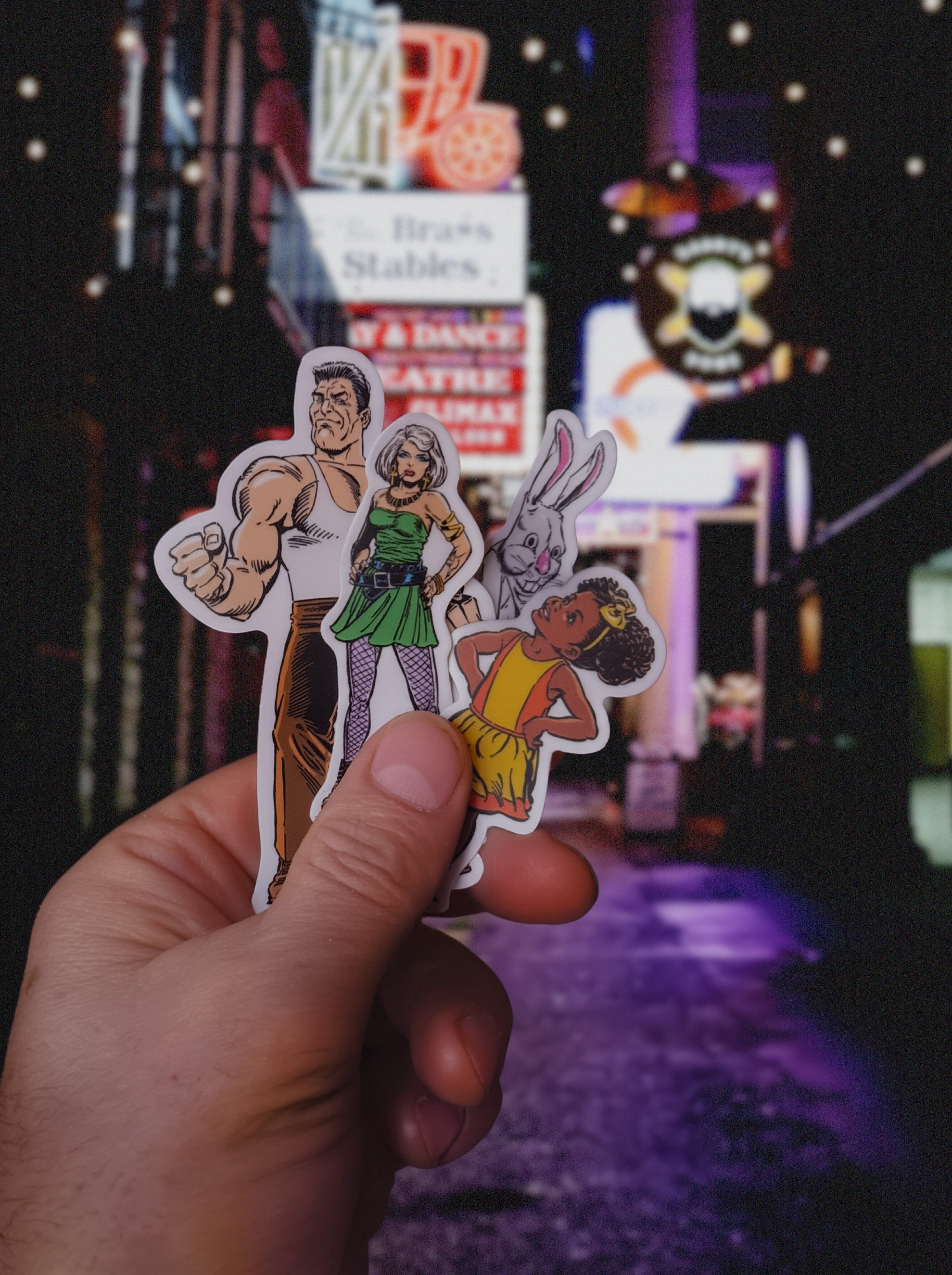 Hand holding a sticker with four animated characters against a dark, neon-lit background.