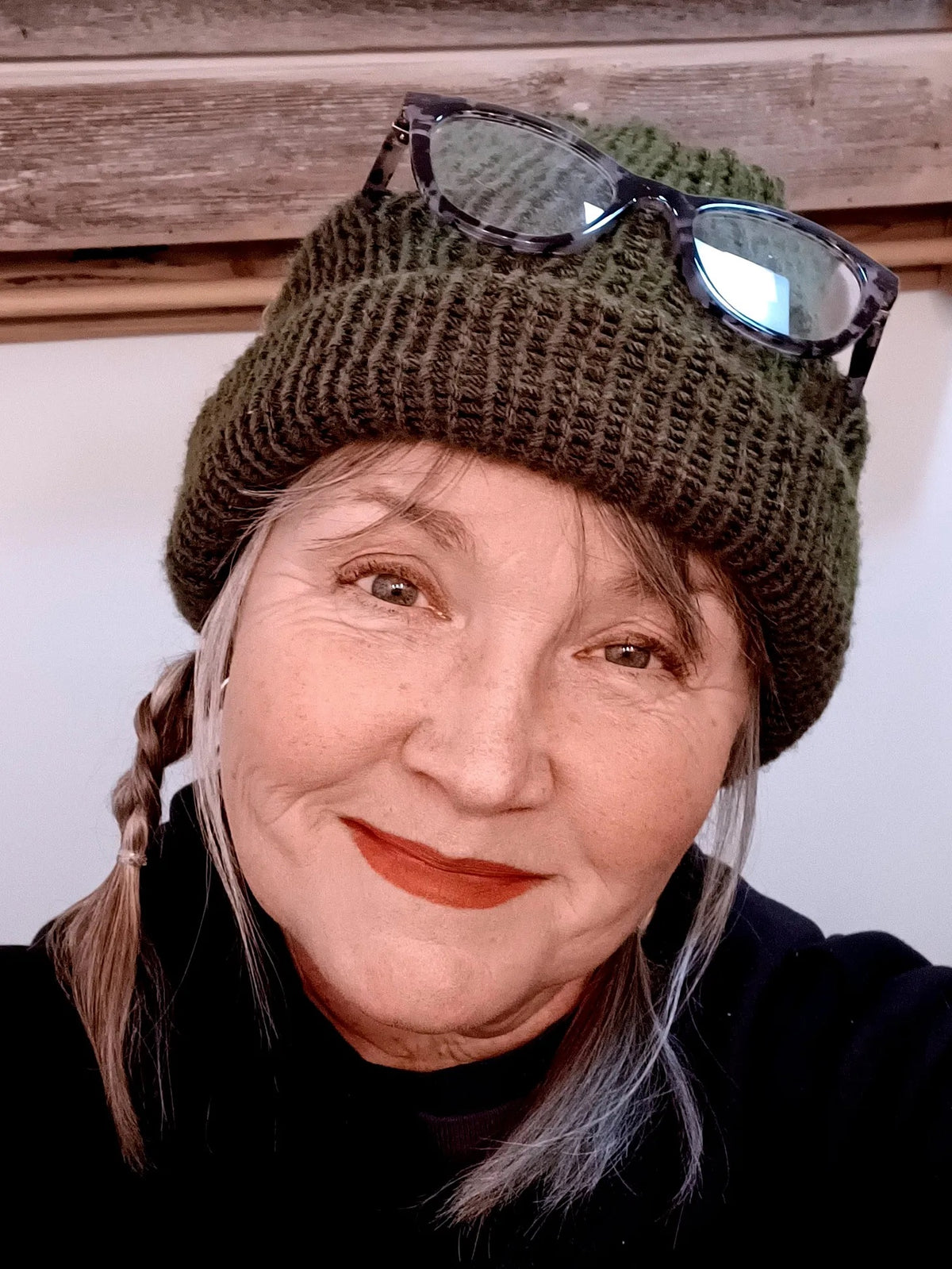 Person wearing a green knit beanie with sunglasses on top, against a wooden panel background.